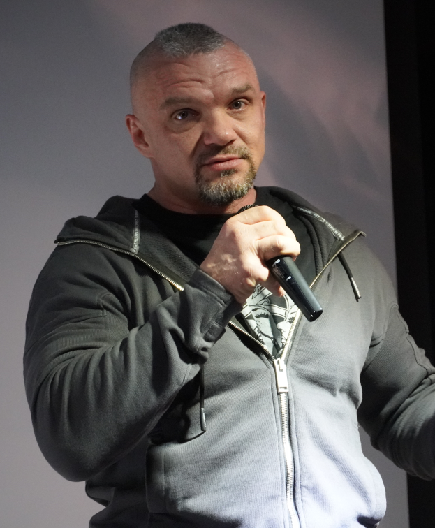 Ein Mann in schwarzer Kapuzenjacke, schwarzem Anzug und weißem Hemd, mit zurückgekämmtem Haar, hält ein Mikrofon und steht selbstbewusst vor einer Wand.