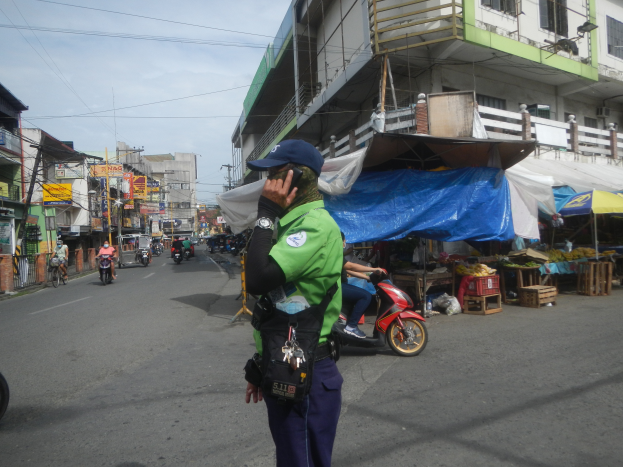 Ein Polizist in grüner und blauer Uniform und blauer Mütze steht an der Straße und spricht in ein Handy, während Fahrzeuge hinter ihm fahren, Stände rechts von ihm und ein bewölkter Himmel über einem Gebäude mit Fenstern im Hintergrund.
