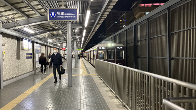 Ein Zug fährt auf einem Bahnsteig ein, Menschen gehen in der Nähe, ein Geländer auf der rechten Seite, Gebäude im Hintergrund, Bahnsteiglampen und ein Anzeigebrett.