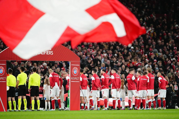 Gruppe von Menschen auf einem Fußballfeld mit einer roten und weißen Flagge im Vordergrund, einem Bogen mit "Bayern München vs. Bayern München Wetten & Vorschau"-Text im Hintergrund und einer großen Menge im Stadion.