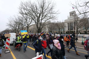 Eine große Gruppe von Menschen nimmt an einer Protestdemo auf einer Straße in Washington, D.C. teil, wobei sie Schilder und Banner schwenken und einige Fahrräder fahren, unter einem klaren blauen Himmel.