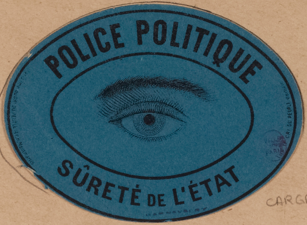 Eine braune Kartonschachtel mit einem blauen Aufkleber, auf dem "Polizei Politique Sûreté de l'État" steht und ein Auge in der Mitte.