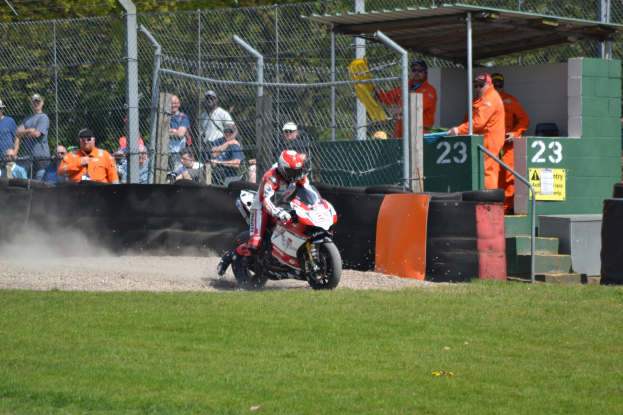 Ein Motorradfahrer mit Helm und Handschuhen fährt über ein grasbewachsenes Feld, mit Zuschauern in Kappen und Schutzbrillen, einem eingezäunten Bereich, Treppen mit Geländern und einem nummerierten Schuppen im Hintergrund.