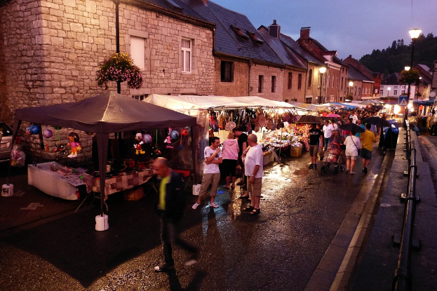 Eine Gruppe von Menschen, die auf der Straße gehen, einige mit Schirmen, mit Läden, die Spielzeug, Taschen und Gebäude mit Fenstern im Hintergrund zeigen, sowie Bäume und Straßenlaternen und Barrieren am Straßenrand.