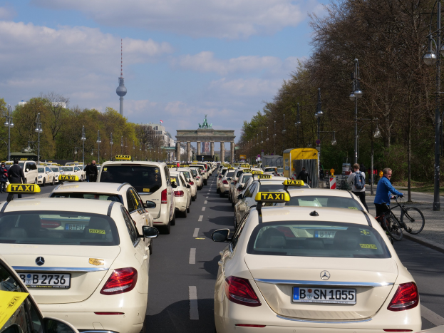 Lange Taxischlange an einer belebten Straße in Berlin, Deutschland, mit Fahrradfahrern und Passanten auf dem Gehweg, flankiert von Laternenmästen und Bäumen, sowie Gebäuden, einem Bogen und einem Turm im bewölkten Himmelhintergrund.