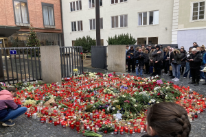 Eine Gruppe von Menschen an einer improvisierten Gedenkstätte mit Blumen, Kerzen und Gegenständen auf dem Boden; eine Person kniet links und hält eine Kamera, mit Gebäuden, Pflanzen, einem Pfahl und einem Tor im Hintergrund.
