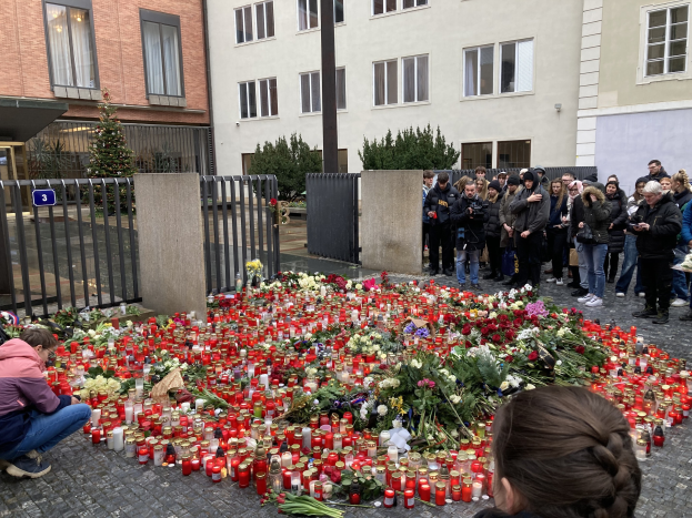 Eine Gruppe von Menschen an einer improvisierten Gedenkstätte mit Blumen, Kerzen und Gegenständen auf dem Boden; eine Person kniet links und hält eine Kamera, mit Gebäuden, Pflanzen, einem Pfahl und einem Tor im Hintergrund.