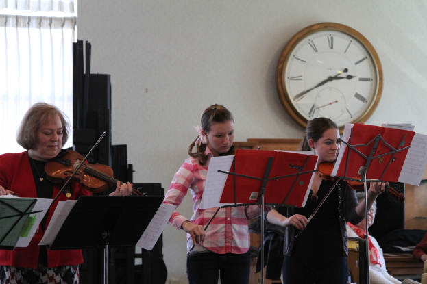Drei Frauen auf der Bühne mit Violinen stehend und spielend, mit Notenständern und Noten davor, während ein Zuschauer im Vordergrund sitzt; eine Uhr ist an der weißen Wand im Hintergrund montiert und ein weißer Vorhang ist links sichtbar.