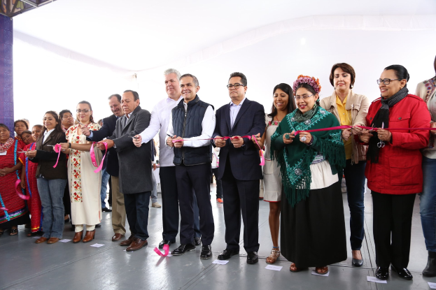 Eine Gruppe von Menschen hält eine Schere bei einer Eröffnungszeremonie vor einer Menge, mit einer Säule links und Papieren auf dem Boden.