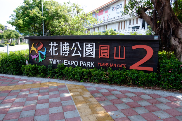 Der Eingang zum Taipei Expo Park 2, mit einem Schild, das von Grün umgeben ist, mit Gebäuden und einem klaren blauen Himmel im Hintergrund.