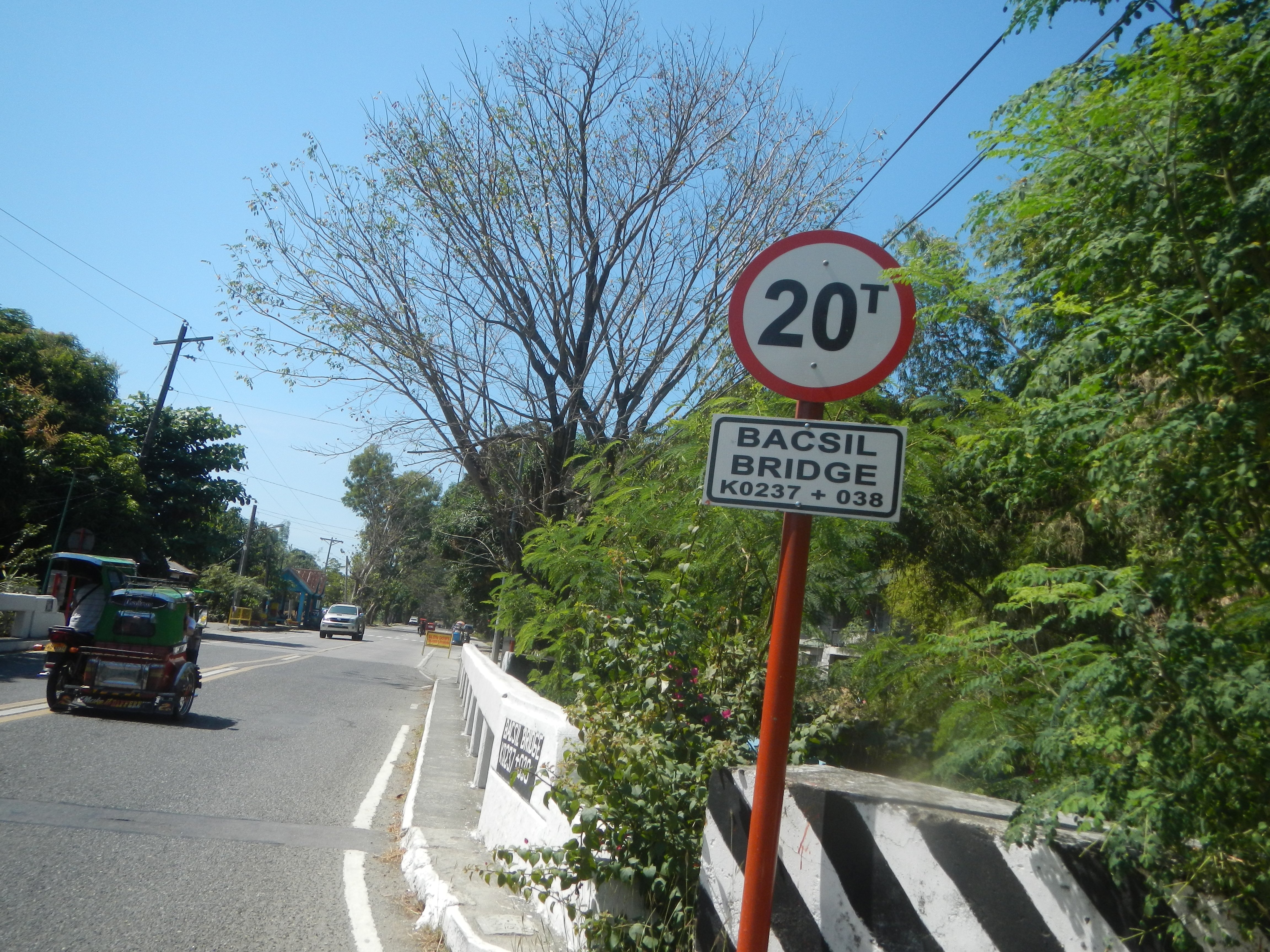 Eine Straße mit Fahrzeugen, ein Tempolimitschild, eine Trennung, Bäume, Strommasten mit Drähten und einen klaren blauen Himmel.