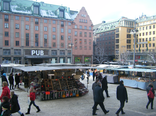 Ein umtriebiger Weihnachtsmarkt in Stockholm, Schweden, mit Menschen, die Görbeutel tragen und Ständen voller verschiedener Gegenstände, vor einem Hintergrund aus Gebäuden, Laternenmasten, Bäumen und einem klaren blauen Himmel.