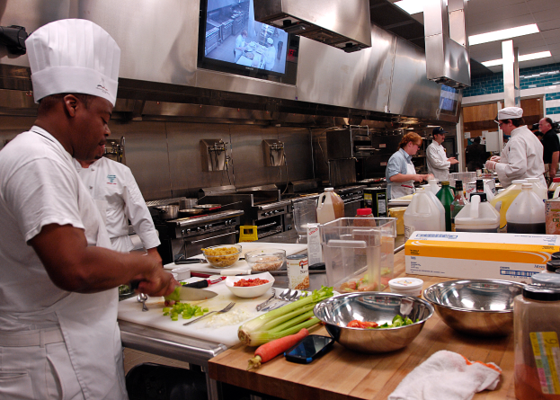 Eine Gruppe von Köchen in einer Küche bei der Zubereitung von Essen auf einem Schneidbrett, mit einem Koch im Vordergrund in einer weißen Schürze und Mütze, der ein Messer hält und von Utensilien, Schüsseln, Gemüse, einem Handy, Flaschen und anderen Gegenständen umgeben ist, mit einem Fernseher und Deckenlampen im Hintergrund.