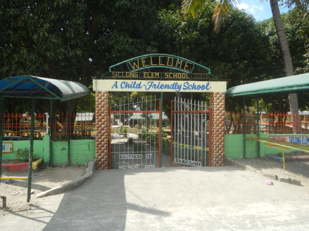 Der Eingang einer kinderfreundlichen Schule mit einem "Willkommen"-Schild oben auf dem Bogen, zwei Unterständen auf jeder Seite und vielen Bäumen sowie einem klaren blauen Himmel im Hintergrund.