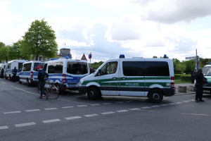 Eine Gruppe von Polizeiwagen, die entlang einer Straße geparkt sind, mit einem Radfahrer im Vordergrund, Bäumen, Pfählen, Schildern, Gebäuden und einem bewölkten Himmel im Hintergrund.