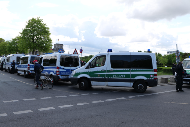 Eine Gruppe von Polizeiwagen, die entlang einer Straße geparkt sind, mit einem Radfahrer im Vordergrund, Bäumen, Pfählen, Schildern, Gebäuden und einem bewölkten Himmel im Hintergrund.