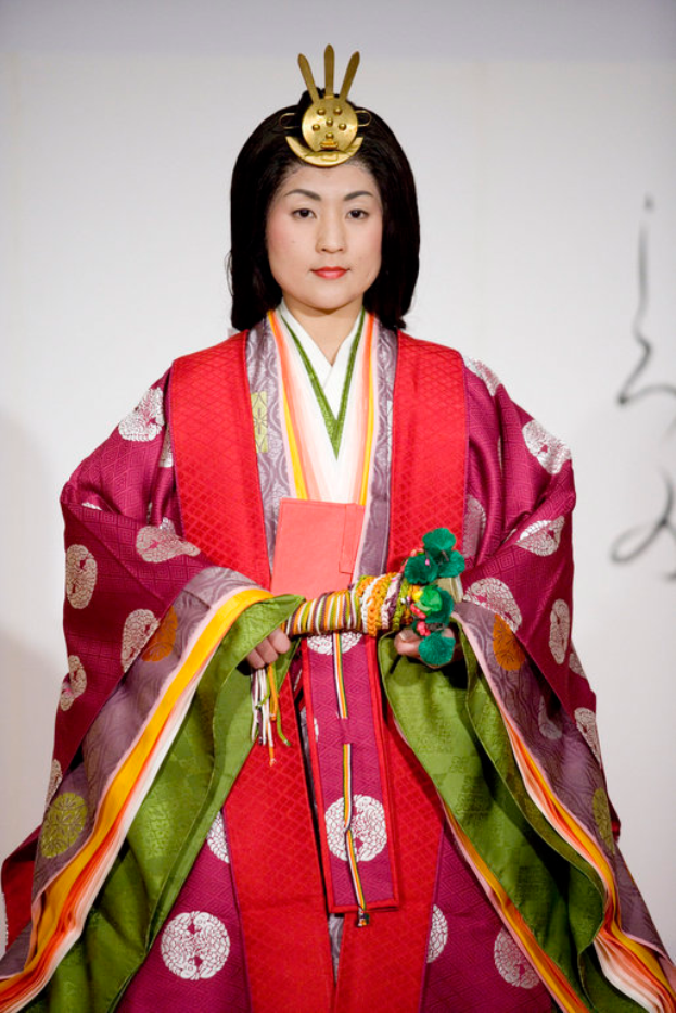 Eine Frau in einem traditionellen japanischen Kimono, das ein leuchtend rotes und grünes Kleid mit aufwändigen Mustern zeigt, steht selbstbewusst auf einem Laufsteg, ihr Haar zu einem ordentlichen Knoten gebunden.