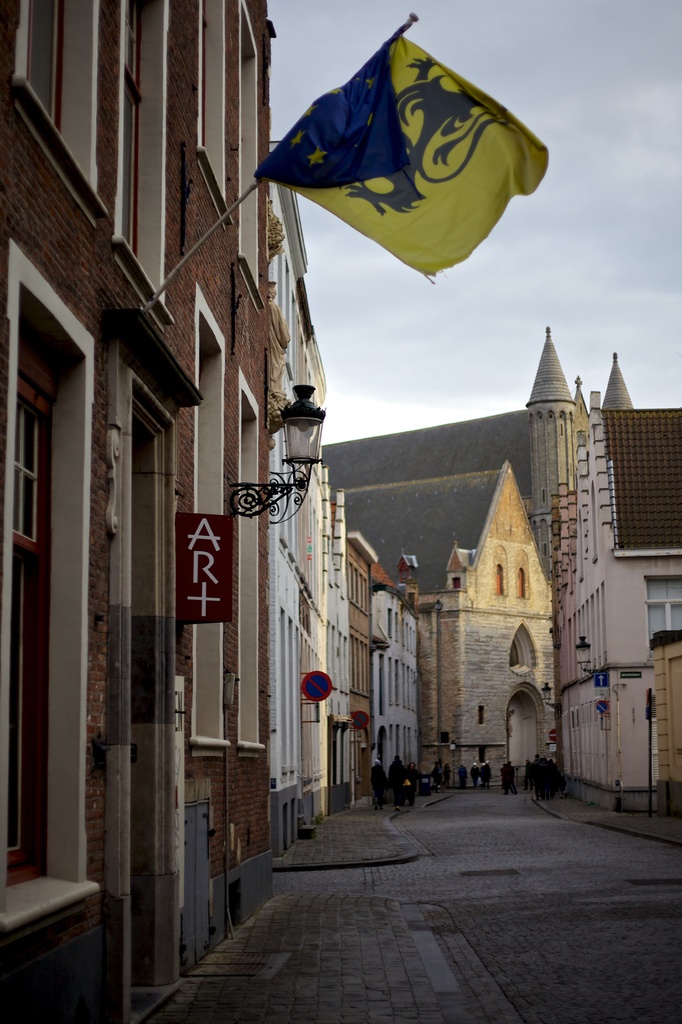 Stadtansicht mit Gebäuden, Menschen auf der Straße, Pfählen, Schildern und einer Flagge an einem der Gebäude, mit dem Himmel oben und der Straße unten.
