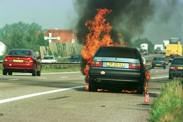 Ein Auto in Flammen an der Straße, umgeben von anderen Fahrzeugen, mit Bäumen, Gebäuden und einem klaren blauen Himmel im Hintergrund und Gras mit einem Feuerlöscher auf der rechten Seite.