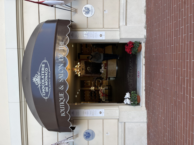 Außenansicht eines Gebäudes mit einer 'Boutique & Salon de Monaco'-Tafel, Außenbestuhlung, Topfpflanzen und einer sichtbaren Inneneinrichtung mit Lichtern und Dekoration.