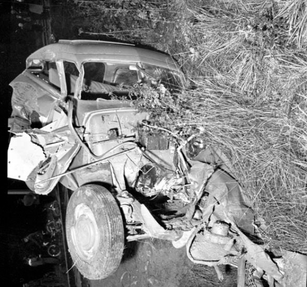 Ein Schwarz-Weiß-Foto eines schwer beschädigten Autos am Straßenrand, mit eingedellter Front, zersplitterten Fenstern, einem Baum im Hintergrund und verstreuten Gegenständen am Tatort.