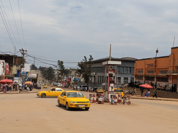 Ein gelbes Taxi fährt eine belebte Stadtstraße mit Gebäuden an den Seiten entlang, während Fußgänger mit Schirmen auf dem Gehweg unter einem bewölkten Himmel gehen, mit Strommasten und Drähten im Hintergrund.