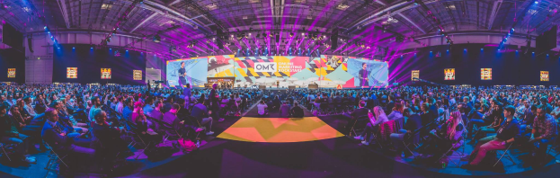 Eine große Menschenmenge, die auf Stühlen sitzt und einer hell erleuchteten Bühne mit mehreren stehenden Individuen zugewandt ist, wahrscheinlich bei einem Online-Marketing-Rockstars-Event, umgeben von Bildschirmen.