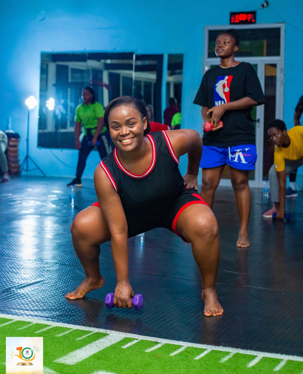 Eine Frau in einem Fitnessstudio führt eine Kniebeuge aus und hält dabei Hanteln, während im Hintergrund verschiedene Fitnessgeräte und Logos zu sehen sind.