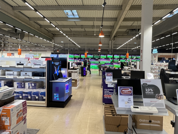 Großer Raum mit Elektronikregalen, die Computer, Drucker und Kartons enthalten, Deckenleuchten und -lüfter, Schilder und Menschen in einem Walmart-Laden.