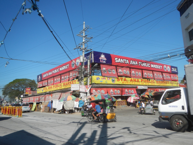 Eine belebte Straße mit einem Lastwagen, mehreren Fahrzeugen, Fußgängern, einem Strommast mit Drähten, Bäumen und einem klaren blauen Himmel.