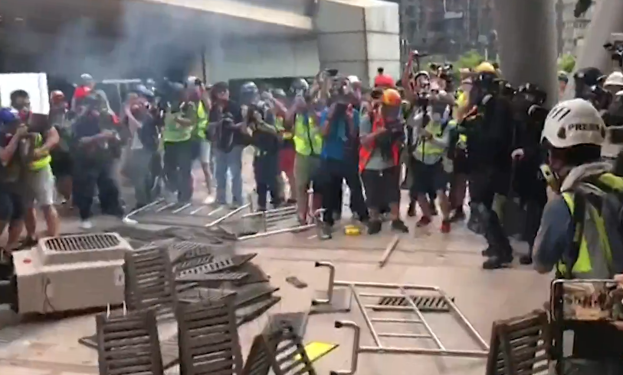 Eine Gruppe von Menschen steht vor einem rauchenden Gebäude, einige tragen Helme und Jacken, andere halten Kameras, mit verstreuten Metallobjekten und Gegenständen auf dem Boden, vor dem Hintergrund von Stadtpfeilern und Gebäuden.