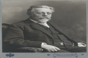 Schwarz-weiß-Fotografie eines bärtigen Mannes mit Brille, der auf einem Stuhl sitzt, mit Text unten, der ihn als "L. Grillich Wien, den Gründer der deutschen Revolution" identifiziert