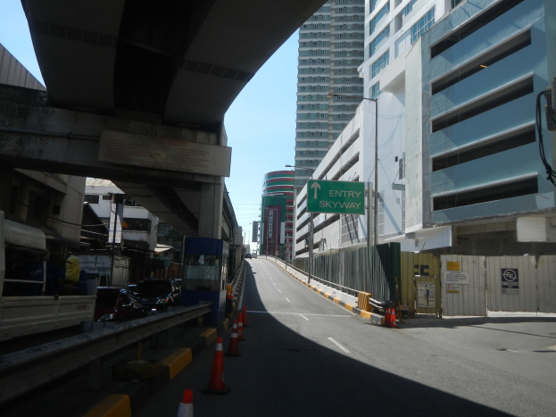 Stadtstraße mit Verkehrskegeln, Fahrzeugen, einer Brücke auf der linken Seite, Gebäuden mit Namensschildern, Laternenmästen und einem Himmel im Hintergrund.