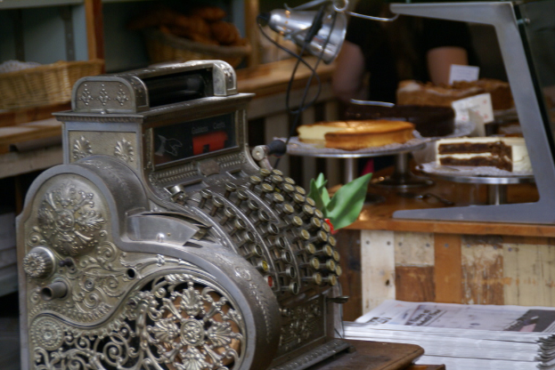 Ein Tisch mit einer Maschine, Büchern, Kuchen auf Ständern, Papieren und einer Lampe, mit Weidenkörben, die verschiedene Gegenstände enthalten, auf einem Schrank in der Nähe.