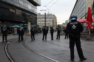 Eine Gruppe von Polizistinnen und Polizisten in schwarzen Uniformen und Helmen steht auf einer von Gebäuden gesäumten Straße mit Glasfenstern und verstreuter Outdoor-Möbel unter einem klaren Himmel.