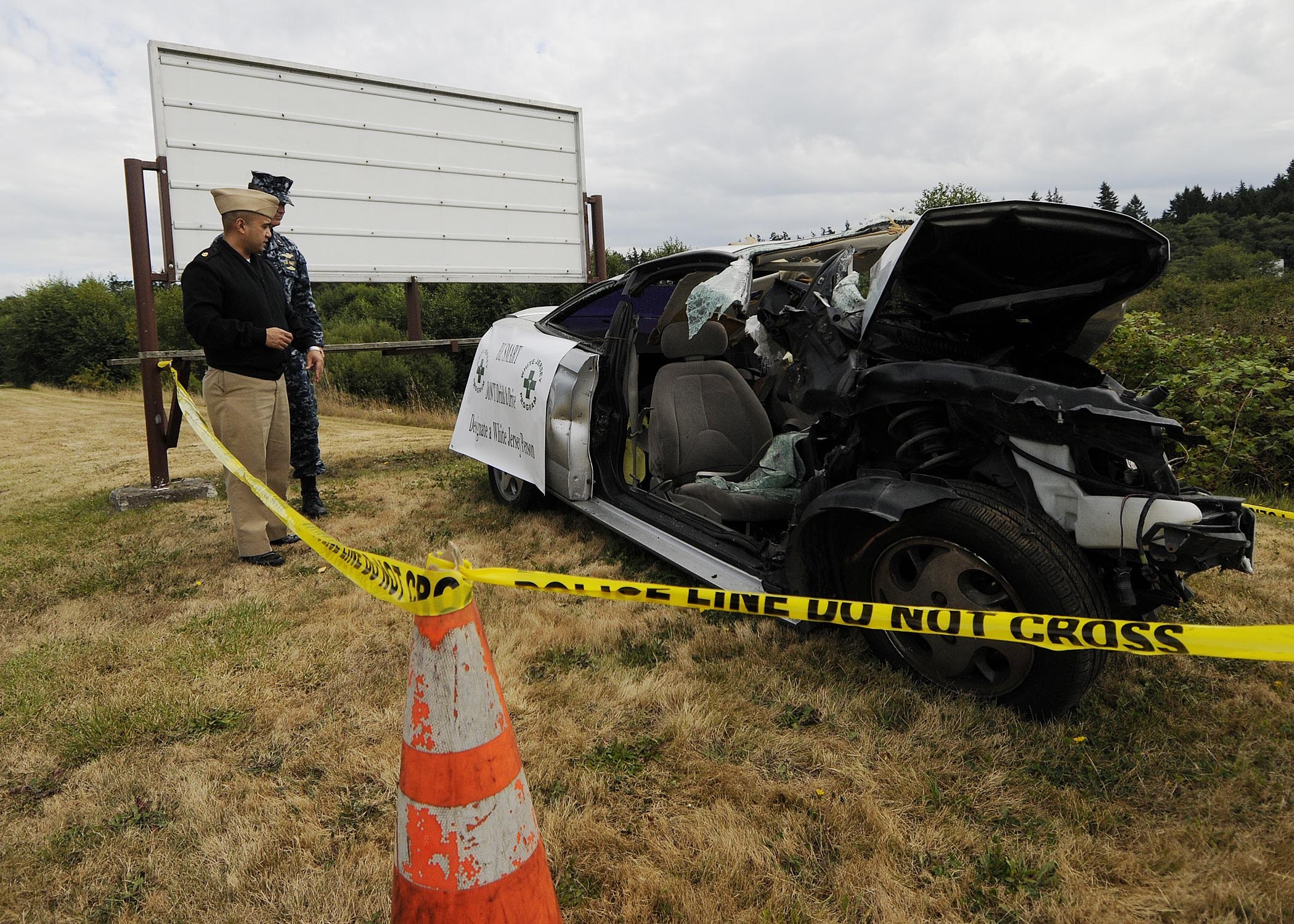 Ein Polizeibeamter in Uniform steht neben einem schwerbeschädigten Auto am Straßenrand mit eingedellter Haube und zersplitterten Fenstern, neben einem Verkehrskegel und einem markierten Pfahl, unter einem bewölktem Himmel mit Bäumen im Hintergrund.