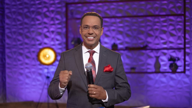 Stephen Colbert, Moderator von *The Daily Show with Trevor Noah*, steht auf einer Bühne und hält ein Mikrofon, trägt einen Anzug, Krawatte und eine Brille, mit einem Scheinwerfer und Ständer im Hintergrund.
