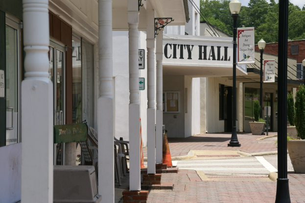 Rathaus mit umliegenden Gebäuden, Straßeninfrastruktur einschließlich Masten, Laternen und Namensschilder, Kraftfahrzeuge, Verkehrskegel, verstreute Stühle, Topfpflanzen, Bäume und ein bewölkter Himmel.