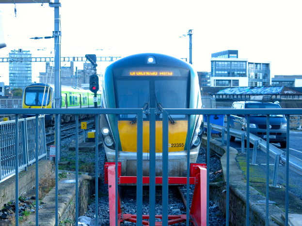 Zwei Züge auf Schienen mit einem Zaun im Vordergrund, Pfosten mit einem mit einer Signalbeleuchtung, ein Auto auf der Straße rechts, Gebäude im Hintergrund und Himmel oben.