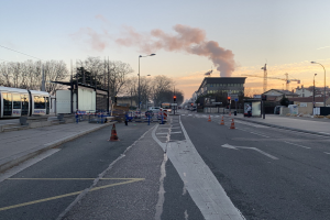 Eine Stadtstraße mit Absperrbaken, einem Bus, Laternen, Bäumen, Gebäuden, Kränen und Rauch, der aus einem fernen Gebäude aufsteigt, vor einem Himmel.