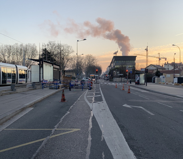 Eine Stadtstraße mit Absperrbaken, einem Bus, Laternen, Bäumen, Gebäuden, Kränen und Rauch, der aus einem fernen Gebäude aufsteigt, vor einem Himmel.