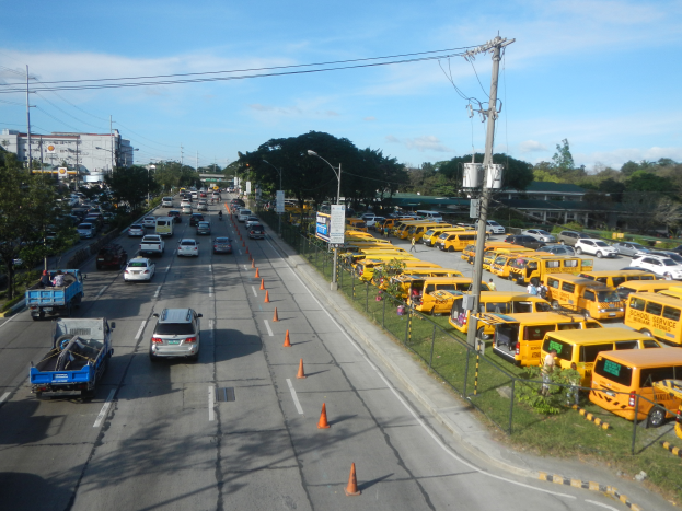 Eine belebte Straße voller Autos, Lastwagen und Bussen, mit Verkehrskegeln, Laternen, Strommästen mit Kabeln und Schildern, einer Maschendrahtzaun auf der rechten Seite und Bäumen, Gebäuden und einem bewölktem Himmel im Hintergrund.