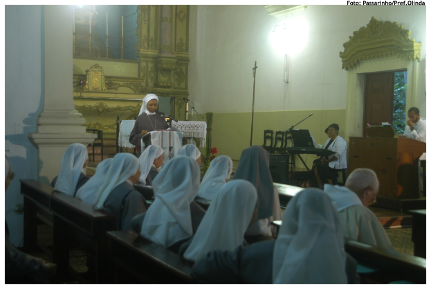 Eine Gruppe von Menschen auf einer Bank, eine Frau an einem Rednerpult, mit Stühlen, mehr Menschen an einem anderen Rednerpult und einer Wand im Hintergrund.