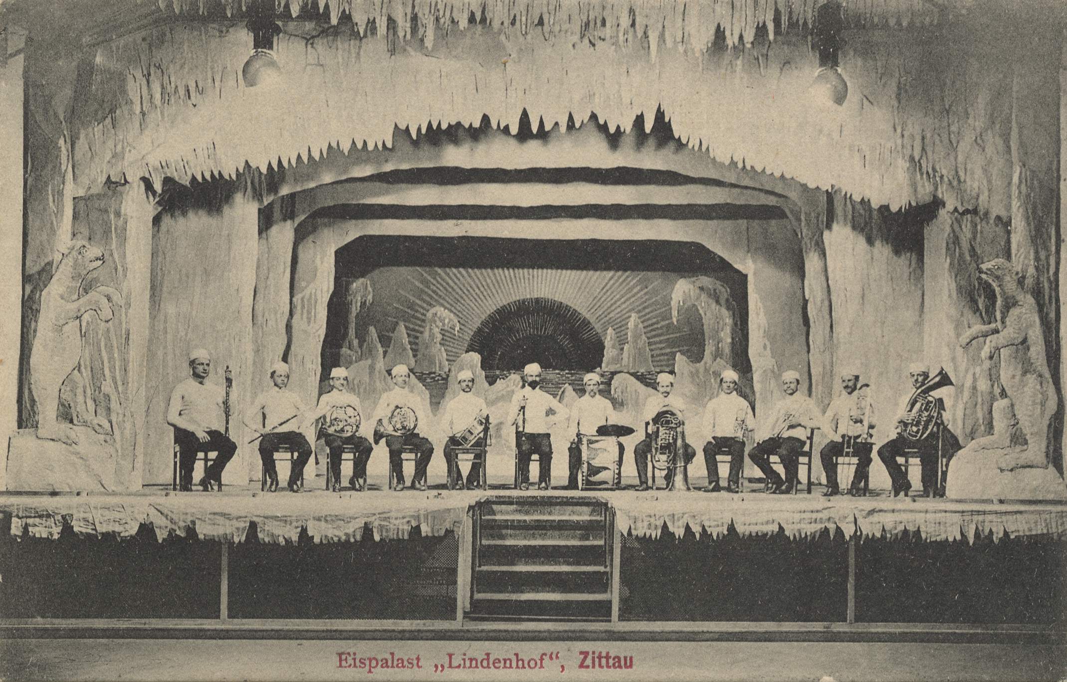 Ein Schwarz-Weiß-Foto einer Gruppe von Menschen auf einer Bühne mit Musikinstrumenten, Treppen, die zur Bühne führen, Skulpturen auf beiden Seiten und Text unten, der 'Lindenhof, Zittau, Deutschland' lautet.