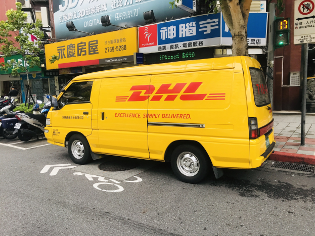 Gelbes DHL-Lieferfahrzeug auf der Straßenseite mit Gebäuden, Bäumen, Schildern, Mästen, Ampeln und einem Fussgänger auf dem Gehweg.