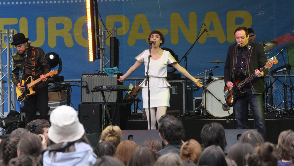 Eine Bühne mit einer Frau in einem weißen Kleid, die in der Mitte singt, zwei Männer mit Gitarren auf der linken und rechten Seite, einer Menge unten und Lautsprechern im Hintergrund.