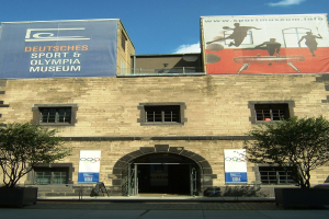 Außenansicht des Deutschen Sport & Olympischen Museums in Berlin, das ein Gebäude mit Fenstern und einer Tür zeigt, umgeben von Bäumen und Bannern unter einem bewölkten Himmel.