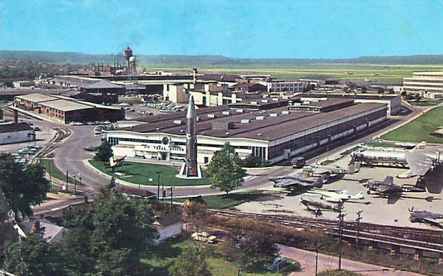 Altes Schwarz-Weiß-Foto eines Flughafens mit parkenden Flugzeugen, Gebäuden, Bäumen, Straßen, Fahrzeugen, Strommasten, Gras, Bahnschienen, Türmen, fernen Hügeln und einem klaren blauen Himmel, mit Text entlang der unteren Kante.