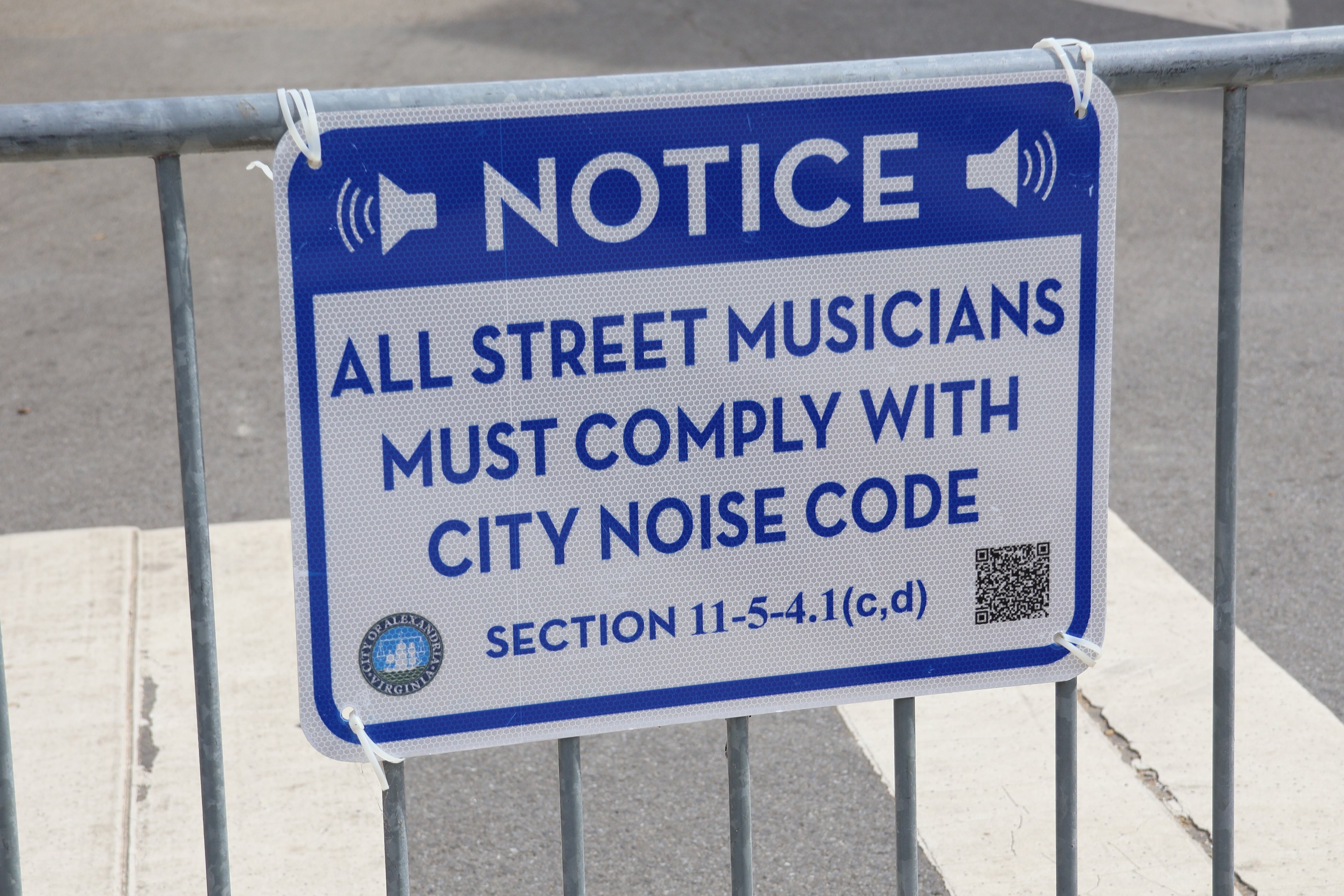 Hinweistafel an einem Zaun mit einem Schild, auf dem steht "Hinweis: Alle Straßenmusiker müssen die städtische Lärmvorschrift einhalten" und eine Straße im Hintergrund.