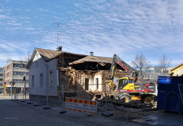 Baustelle mit einem großen Gebäude, das abgerissen wird, mit einem Kran, einem Zaun, einer Absperrung, Steinen, einem Container, Bäumen, Pfählen, einem Schild, einem Straßenpfosten, Fahrzeugen, Gebäuden mit Fenstern und einem bewölkten Himmel.
